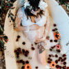 pregnant woman in a milk bath holding her belly with red, orange and pink flowers surrounding her. She is wearing a white lace dress and a flower crown