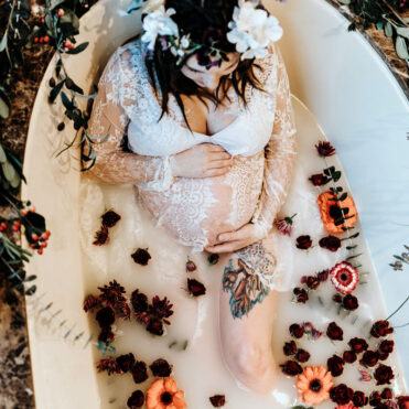pregnant woman in a milk bath holding her belly with red, orange and pink flowers surrounding her. She is wearing a white lace dress and a flower crown