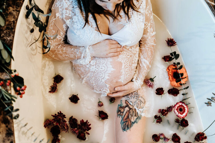 pregnant woman in a milk bath holding her belly with red, orange and pink flowers surrounding her. She is wearing a white lace dress and a flower crown