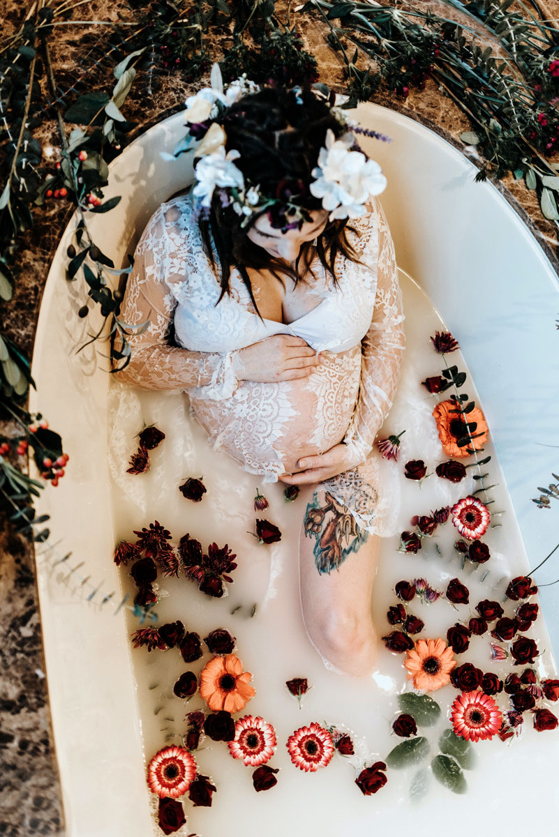 pregnant woman in a milk bath holding her belly with red, orange and pink flowers surrounding her. She is wearing a white lace dress and a flower crown